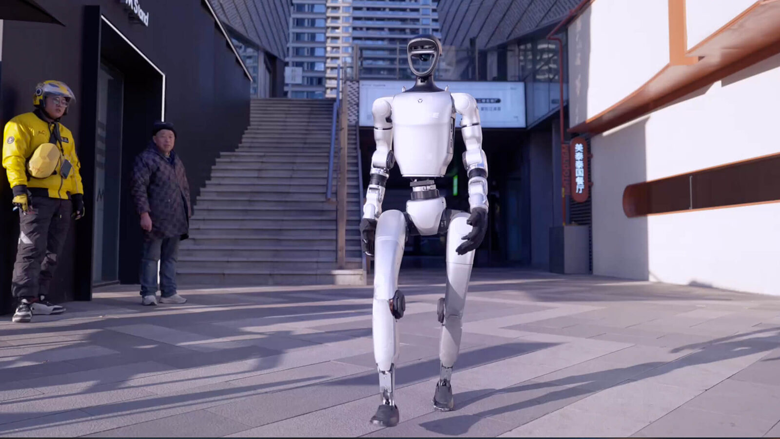 Unitree G1 humanoid robot walking along a street with a staircase behind it
